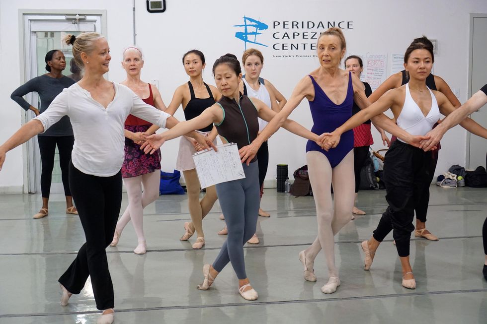 A group of older adult ballet students in leotards, tights or leggings, stand in two lines with their left foot in B+ position and holding hands, as if rehearsing a ballet.