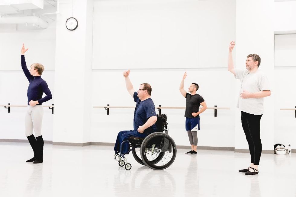 A group of four men in dance practicewear face the right corner of the room and raise their arm as if beckoning someone. Three of the men stand in parallel, which the man in the middle sits in a wheelchair.