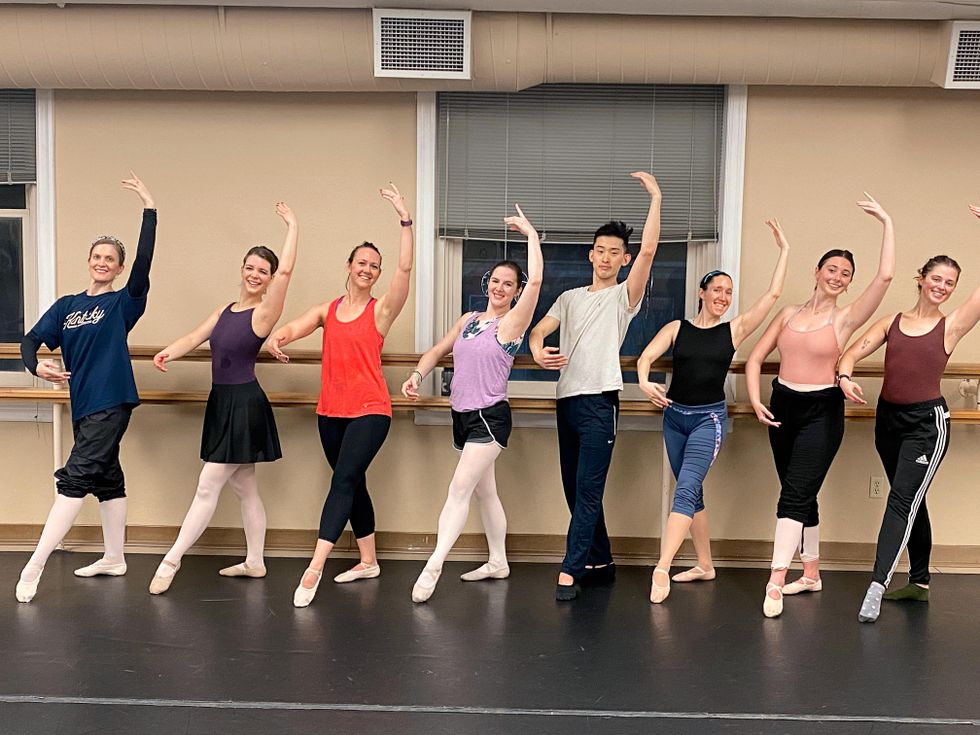 A group of eight smiling adult ballet studentsu2014seven women and one man in the middleu2014pose in a line and stand on their right leg in tendu croisu00e9 devant.