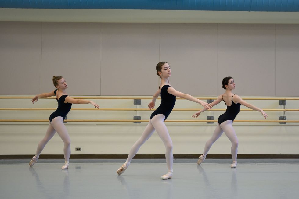 Three ballet dancers take class in black camisole leotards, pink tights and pointe shoes. They are standing in tendu croisu00e9 derriu00e8re, with their left leg in pliu00e9 and their arms in fourth position arabesque.