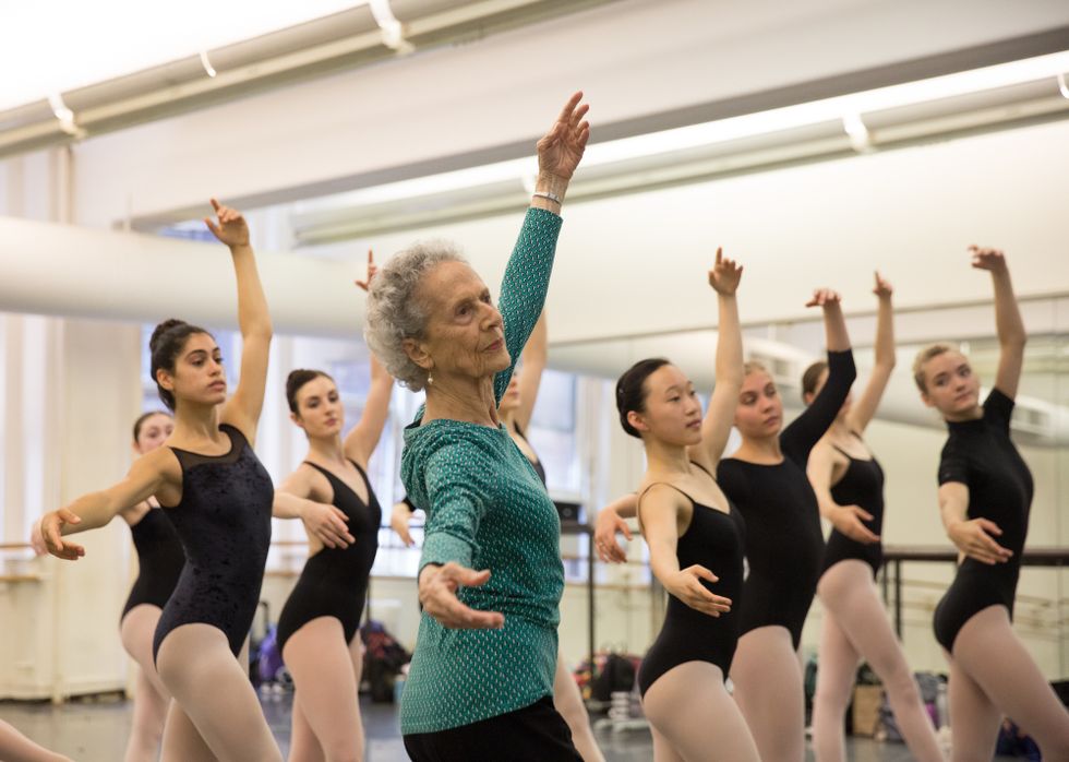 Lupe Serrano, shown from the waist up and wearing a long-sleeved green shirt and black pants, demonstrates tendu croisu00e9 devant. A group of teenage dancers in lack leotards and pink tights stand behind and to the right of her, imitating her position.