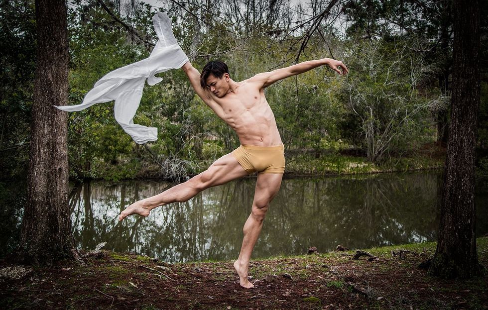 Chun Wai Chan, wearing tan booty shorts and near a wooded creek, piques on his left foot and stretches his right leg in degagu00e9 u00e0 la second. He lifts his right arm up and tosses a gauzy white shirt.