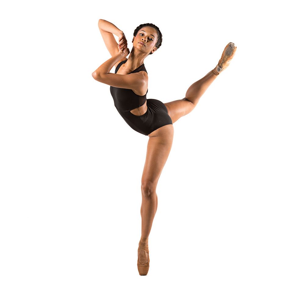 Ho-Tsau00ef in a black leotard poses in a back attitude with her hands gripped in the air against a white background.