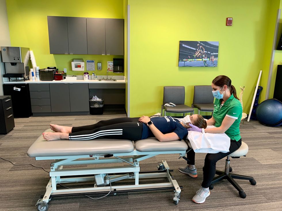 A dancer lies flat on a therapy table while a therapist cradles the dancer's head in her hands to test neck flexibility.