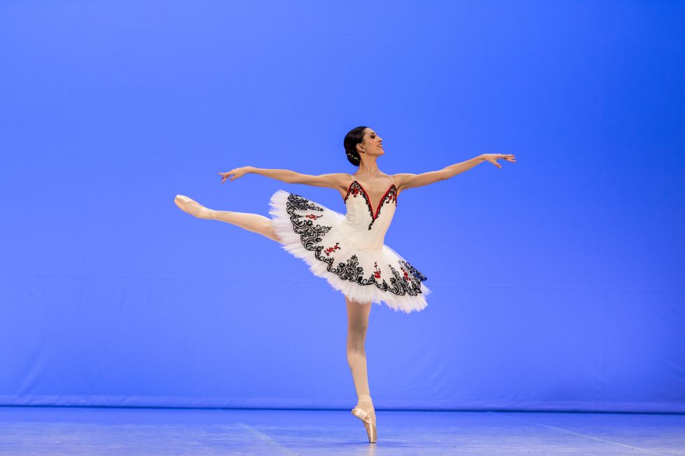 Carolina Pires does a piquu00e9 first arabesque on pointe on her left leg. She smiles brightly, wearing a white tutu with black and red trim, pink tights and pointe shoes. Her dark brown hair is pulled into a low bun.