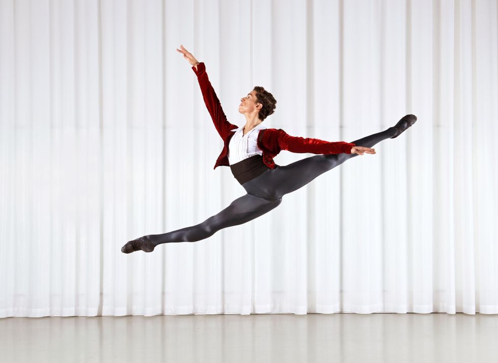 Wearing a red bolero jacket, black tights and cummerbund and a white shirt, Luca Abdel-Nour does a large sissone to the right, his legs in a deep split.