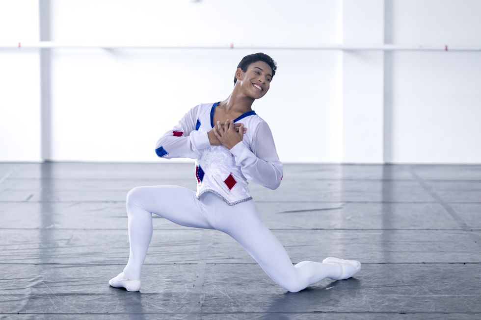 Andrey Jesus Maciano, a Brazilian teenage boy, wears a white Harlequin costume with blue and red diamonds. He kneels onto the floor on his right leg and holds his hands to his heart, smiling wistfully.