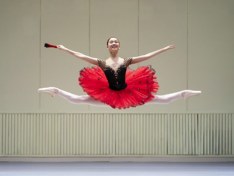 Seojeong Yun, a teenage female ballet student, wears a black and red tutu and does a giant jetu00e9 with both legs split in second position and her arms out to the side. She wears a large, confident smile.