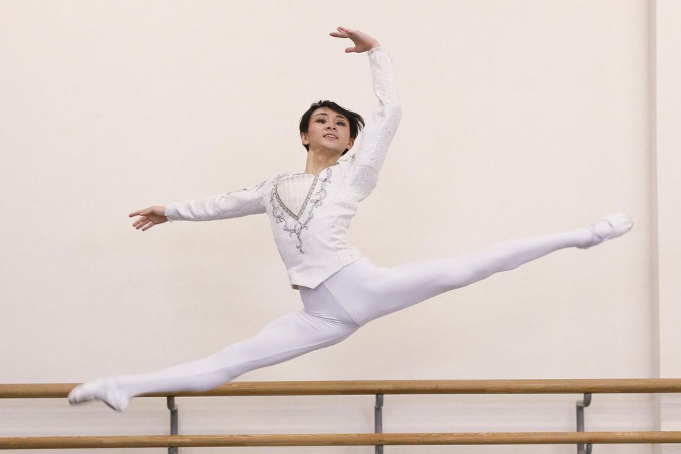 Shunhei Fuchiyama, a teenage Japanese ballet student, wears an all-white cavalier costume and performs a large split sissone to the right. He looks out towards the audience with a smile.