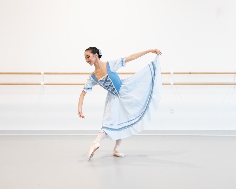 Ashley Coupel, a Canadian ballet dancer of Asian decent, wears a blue and white peasant costume dress and performs a tendu devant effacu00e9. She holds her skirt with her left hand and bends slightly over her curves her right arm, with a shy smile and downward eyes.
