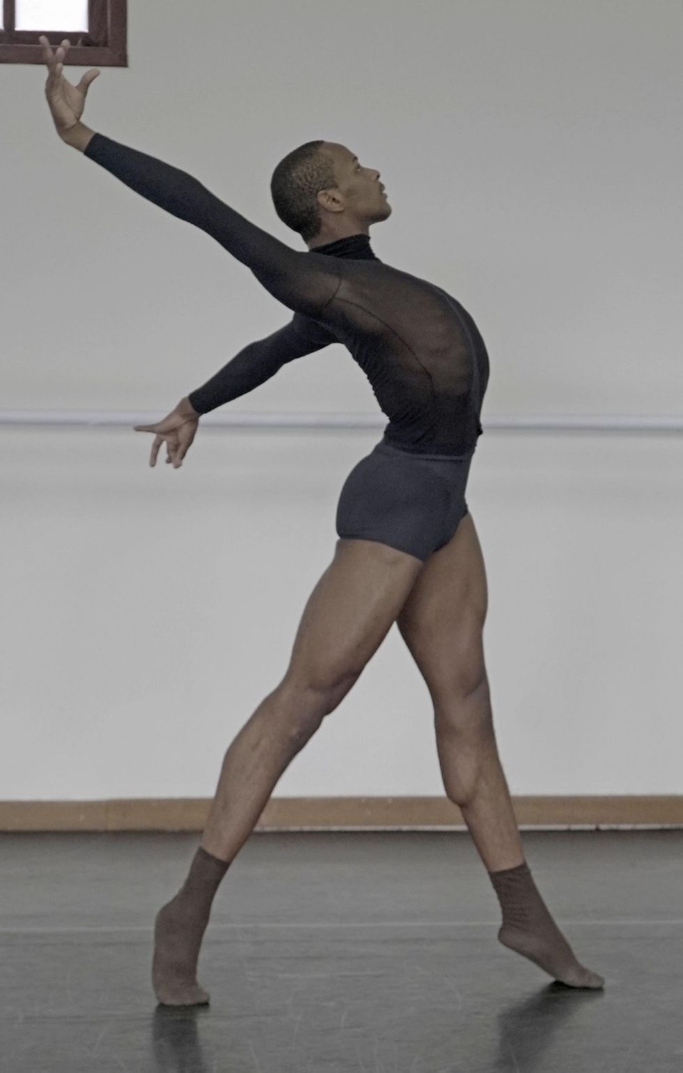 Rui Cesar Cruz, a teenge Afro-Brazilan dancer, does a relevu00e9 effacu00e9 to the left and arches his back dramatically, his arms stretched up and behind him. He wears a black mesh shirt, gray booty shorts and brown socks.