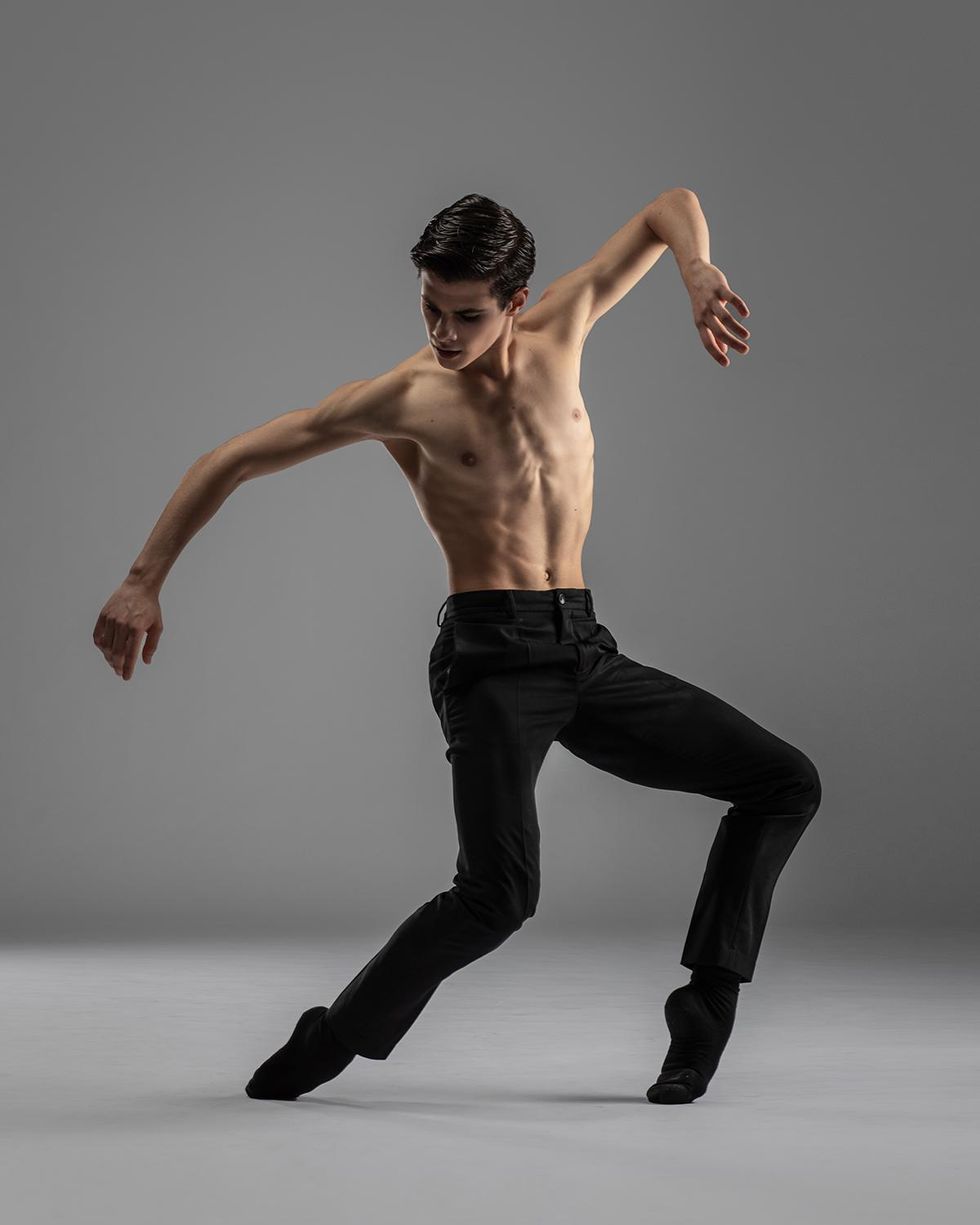 Antonio Casalinho, shirtless and in black jeans , socks and ballet slippers, presses over his toes with two bent knees, lifting his arms up slightly.