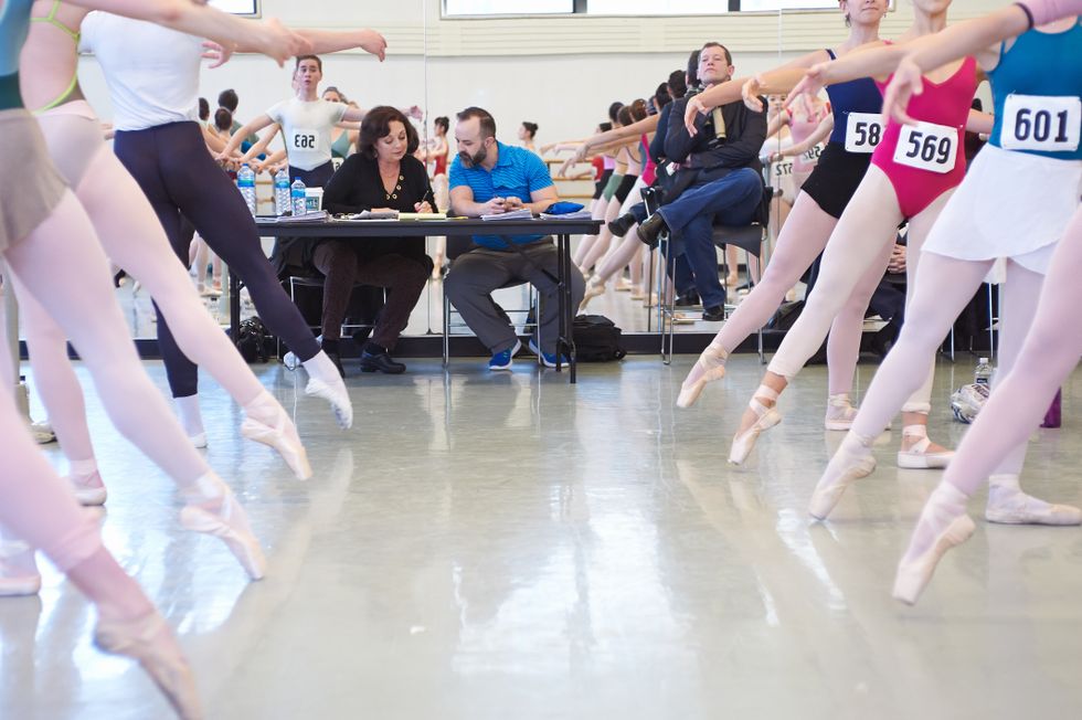 At a table against a studio mirror, a middle-aged man and woman review resumu00e9s and photos while Adam Sklute sits next to them in a chair. Two barres of auditioning dances flank the photo on each side.