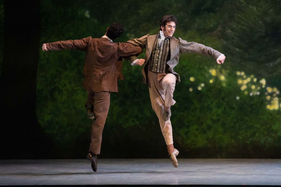 During an onstage performance, two young men in costumes of jackets and pants link arms and skip in front of a leafy backdrop.