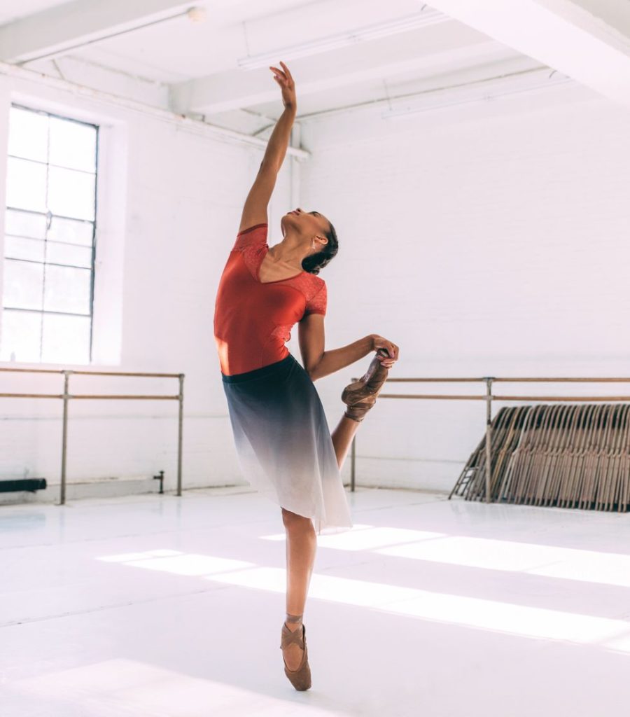 Wearing an orange leotard, blue ombré skirt and pancaked pointe shoes, Smith holds onto her back foot and looks up at one hand in the studio.