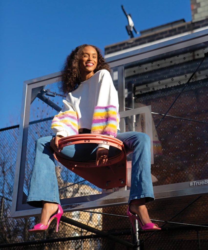 Smith sits suspended in the air on a basketball hoop wearing a fuzzy sweater, bellbottom jeans and pink heels.