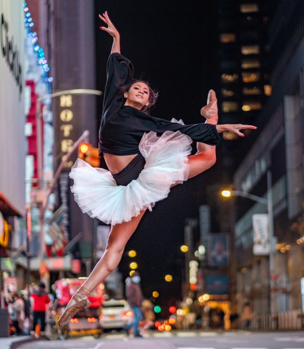 In New York's Times Square, an African American ballerina in a black long-sleeved shirt and white tutu performs a large jetu00e9 with her left leg bent and nearly kicking her head.