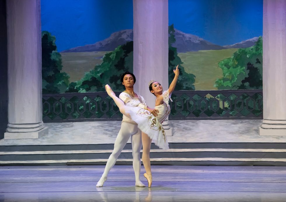 A ballerina wearing a white tutu poses in a high attitude effacu00e9 on pointe during a performance. Her male partner, wearing a white jacket and tights, stands behind her in tendu, holding her waist.