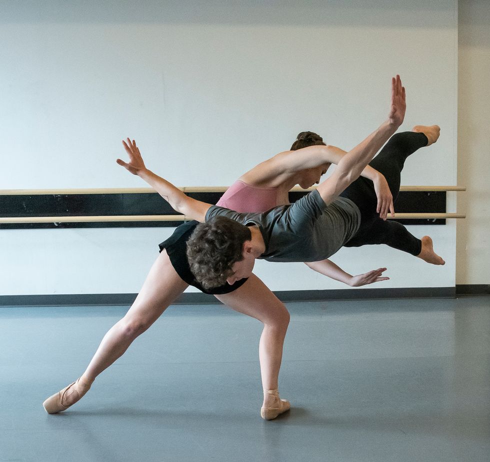 A female dancer lunges in tendu derriu00e9re, making a circular shape with her arms. A male dancer jumps through her arms, his body horizontal in the air and his own arms outstretched.