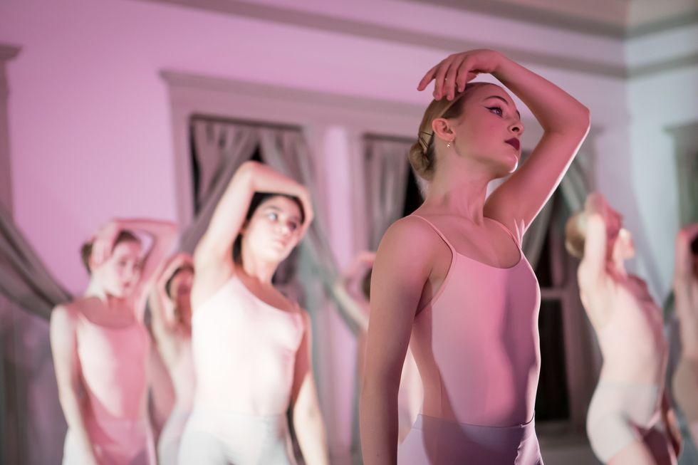 A group of teenage ballerinas in pink leotards wrap one arm around their head during an in-studio performance.