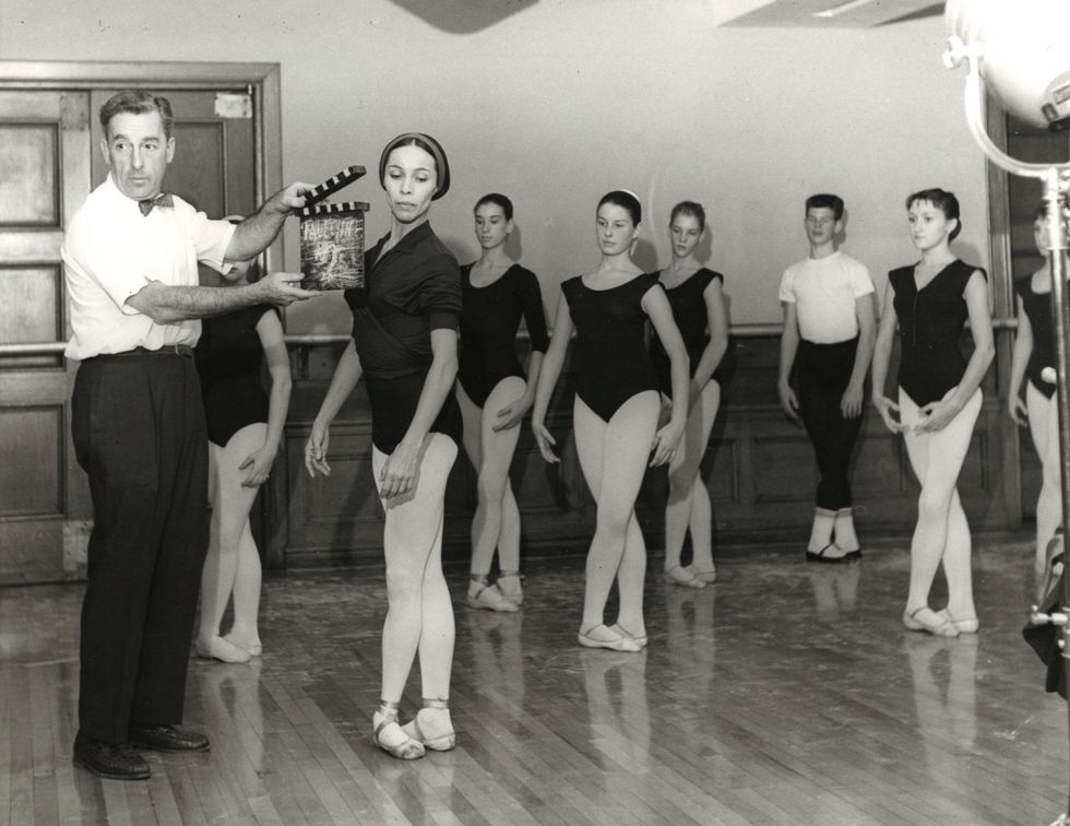 In this black and white photo from the 1960s, a group of female dancers stand in fifth position wearing black leotards, tights and pointe shoes. A man in a white short-sleeved shirt, bow tie and black pants stands to the left holding a clapperboard.