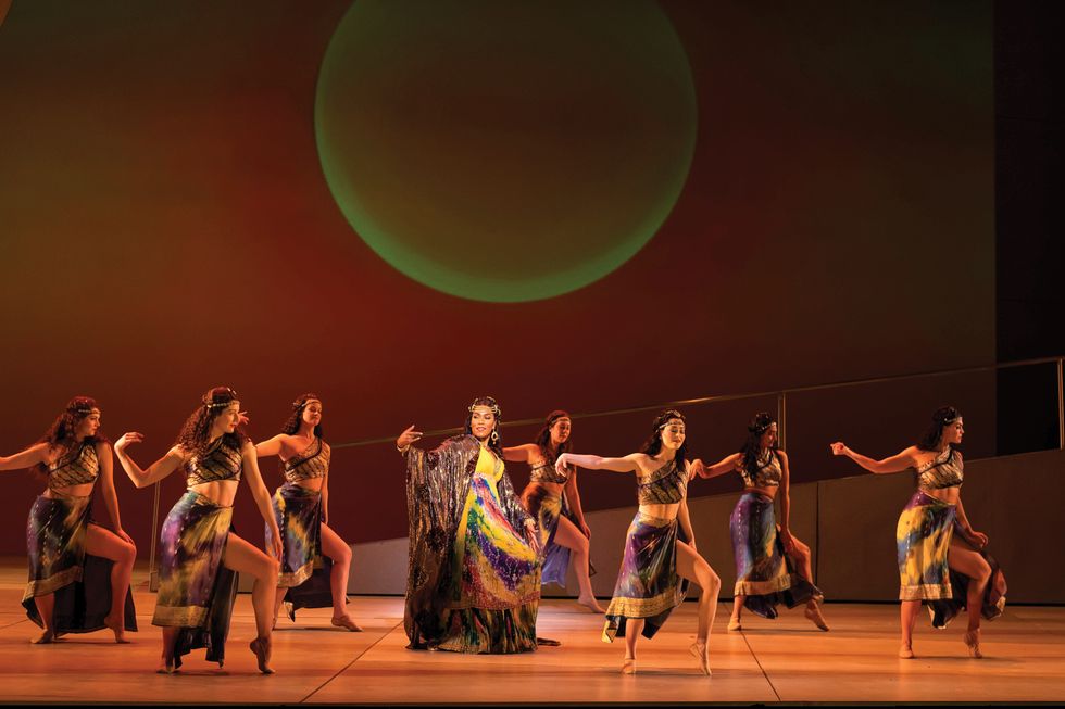 A group of dancers stand with one hip popped onstage wearing tie dye wrap skirts and gold bodice tops with a singer in the middle.