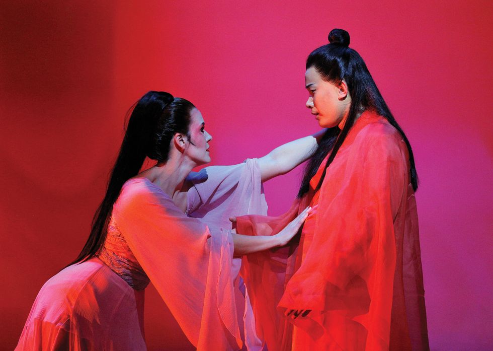 Two dancers dressed in red robes with long black hair against a red background. Little leans over and presses on the actor's chest.