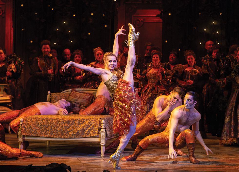 Campbell, lying on a brocade gold bed onstage, while Mearns in gold heeled boots and a gold and red costume stands with her leg kicked high in the air in front of her.