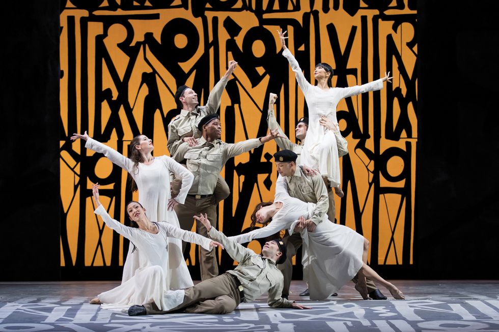 A group of dancers in white dresses and military outfits pose onstage in front of a yellow and black geometric background.