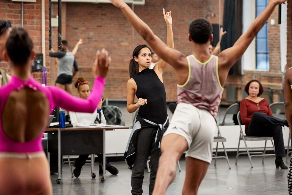 Inside a brick-walled dance studio, choreographer Claudia Schrier wears dance clothes and points upward to demonstrate what she wants to a room of dancers.