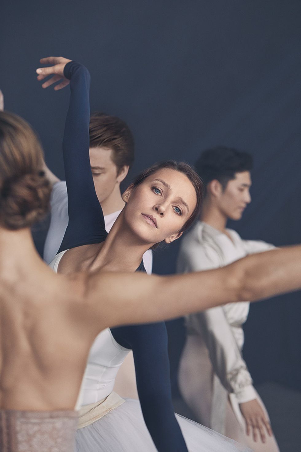 Devon Teuscher looks into the camera. There is another dancer, facing backwards, in the foreground, and two male dancers in the background.