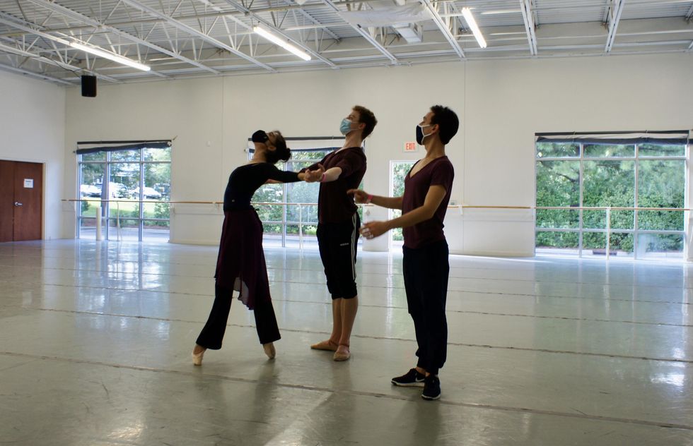 In a large dance studio, Courtney Schenberger, wearing a black leotard, skirt face mask and wide legwarmers, stands in fourth position on pointe with her arms extended behind her. Her partner, Sam Ainley, holds onto her hands and stands in a casual first position. Pablo Perez stands to their left and gestures as if giving them a note.