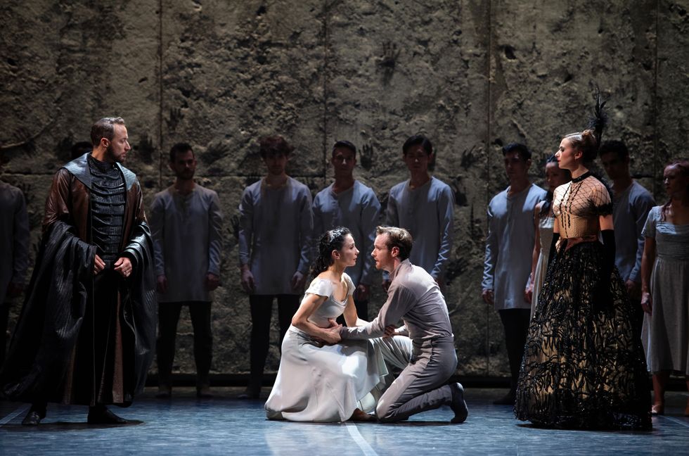 Tamara Rojo, in a white dance dress, and James Streeter, wearing a gray shirt and pants, face each other and crouch down, holding hands. A group of costumed dancers surround them in a half circle.