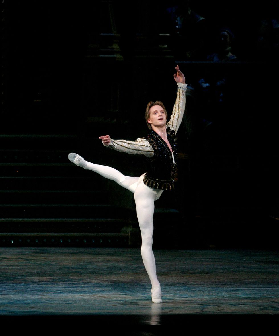A male dancer in white tights and ballet slippers and black embellished tunic does piquu00e9 attitude derriere croisu00e9 on his right leg onstage.