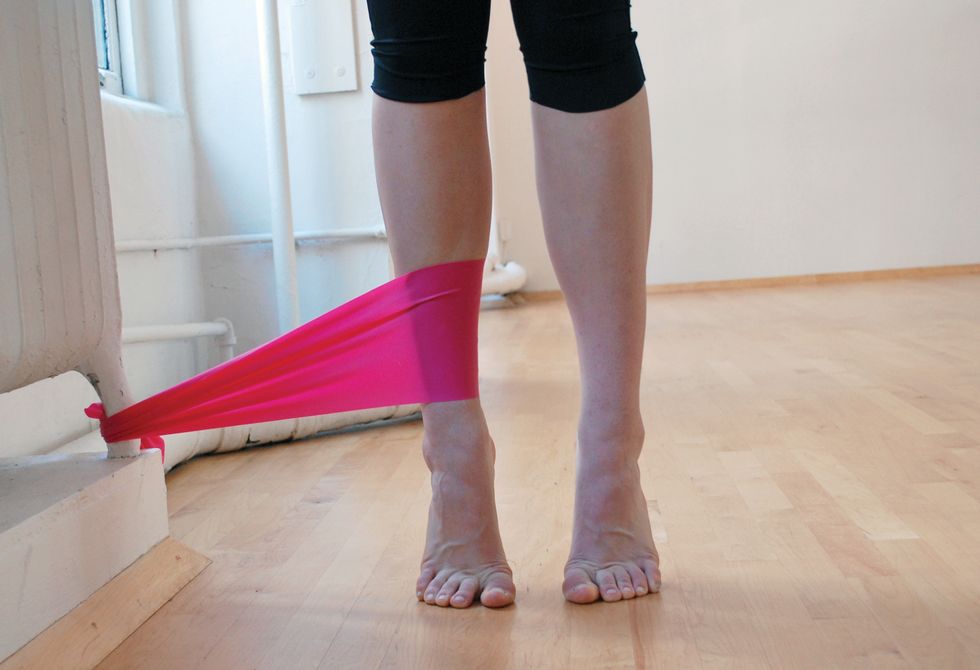 A dancer stands in black leggings on a wooden studio floor. We only see her legs. She is standing in relevu00e9, and a pink elastic band attaches her ankle to the base of a white radiator.