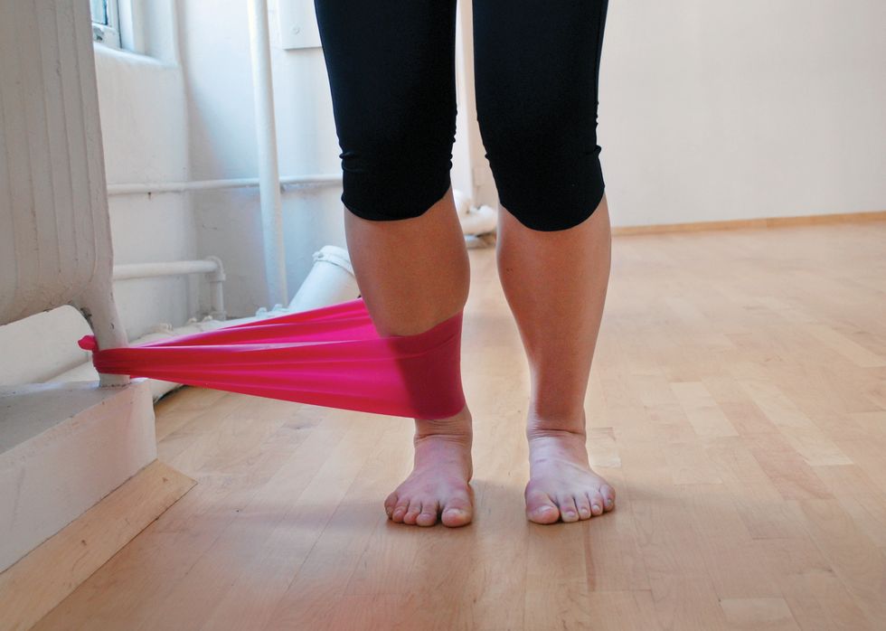 A dancer stands in black leggings on a wooden studio floor. We only see her legs. She is standing in pliu00e9, and a pink elastic band attaches her ankle to the base of a white radiator.