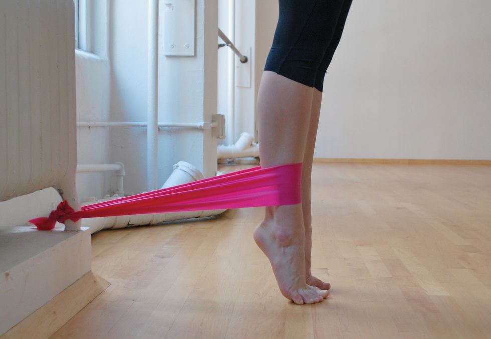 A dancer stands in black leggings on a wooden studio floor. We only see her legs. She is standing in relevu00e9, and a pink elastic band attaches her ankle to the base of a white radiator. We see her legs in profile..