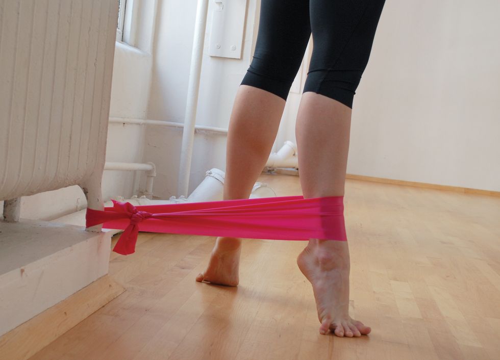 A dancer stands in black leggings on a wooden studio floor. We only see her legs. She is standing in relevu00e9 turned out, and a pink elastic band attaches her ankle to the base of a white radiator.