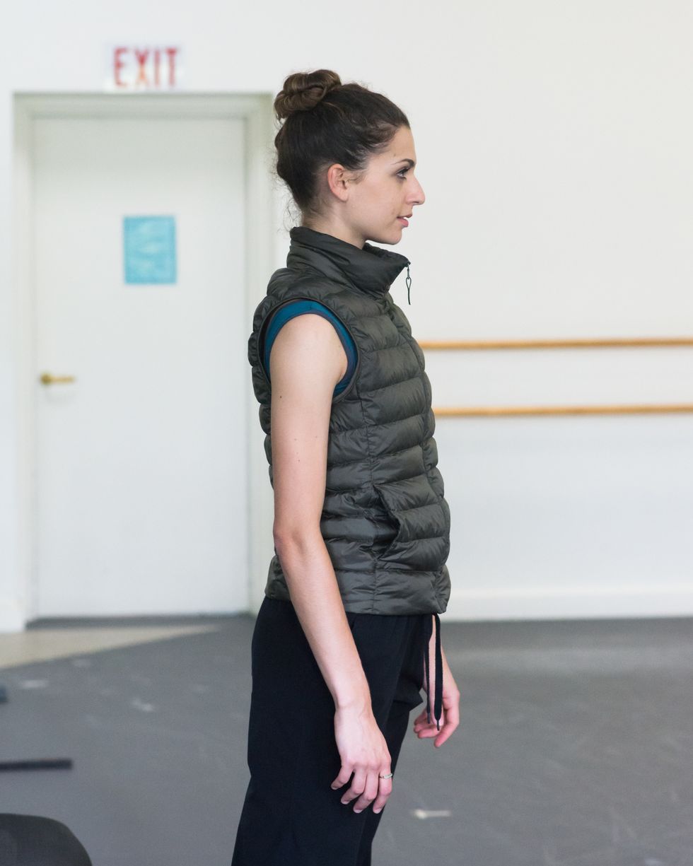 Lauren Flower stands in profile, arms by her side, wearing a dark green puffy verst and black sweat
