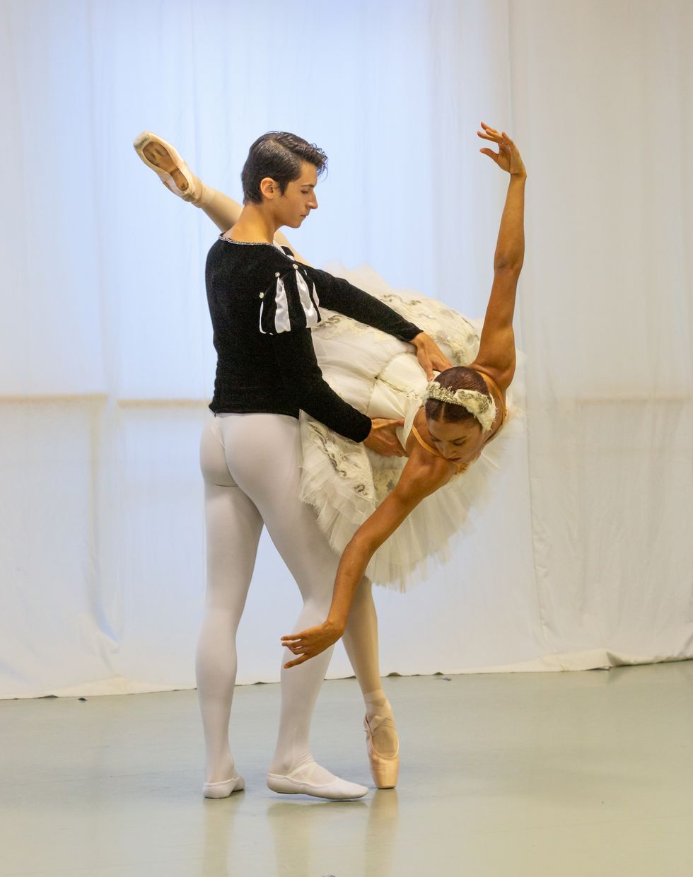 A young male dancer in white tights and a dark velvet dance tunic holds the waist of a female ballerina in a white swan tutu as she takes penchu00e9 on pointe.