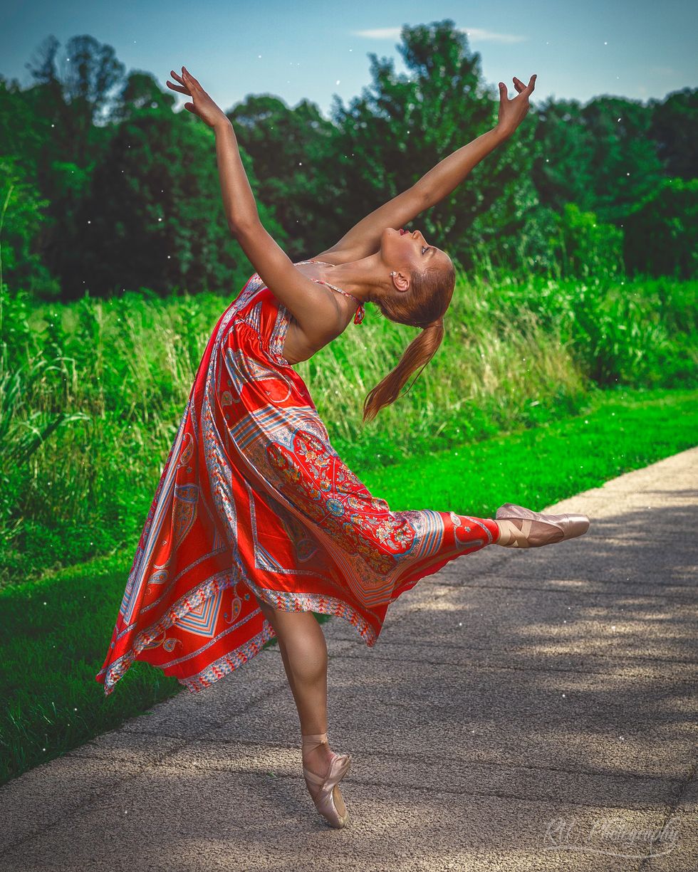 Ballerina Audrey Malek, wearing a red paisley dress and pointe shoes, stands on pointe in a low attitude, her arms in third arabesque position, and leans back in her upper body.