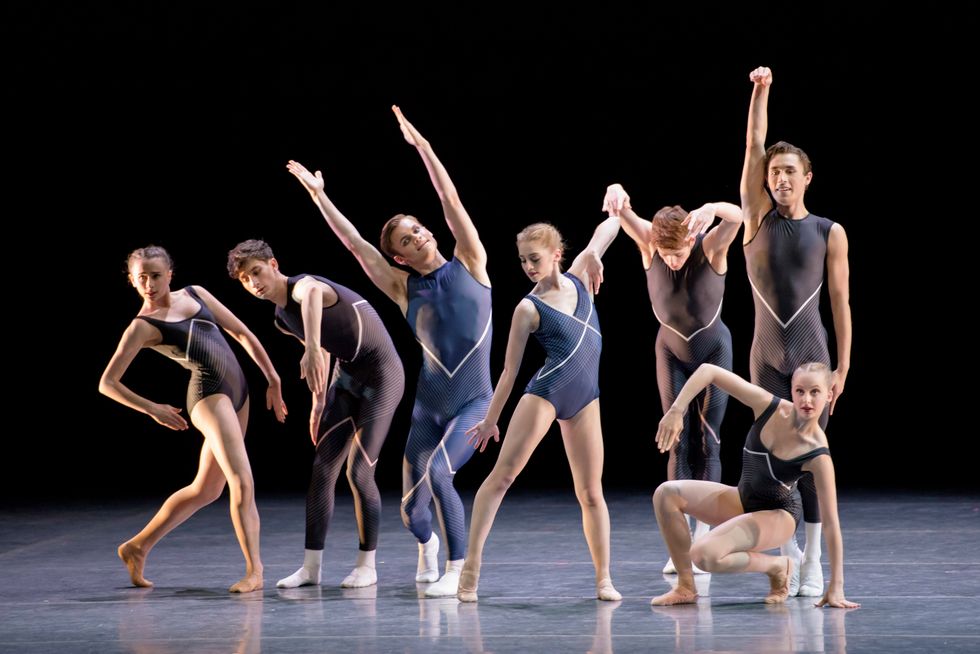 A group of three girls and four boys in blue and black leotards and unitards pose in various modern shapes onstage in front of a black background.