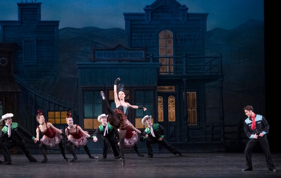 Lauren Ostrander wears a dark tutu, black tights and pointe shoes, and a blue feather on her head and kicks her leg in developpu00e9 ecartu00e9. Behind her is a line of dancers dressed as saloon girls and cowboys, and a lone cowboy stands on the right side of the stage looking on.