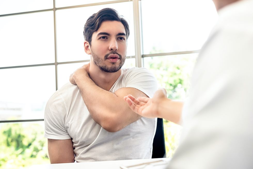 A young athletic man with a short, dark beard and white V-neck T-shirt clutches his right shoulder with his left hand and talks to a doctor.