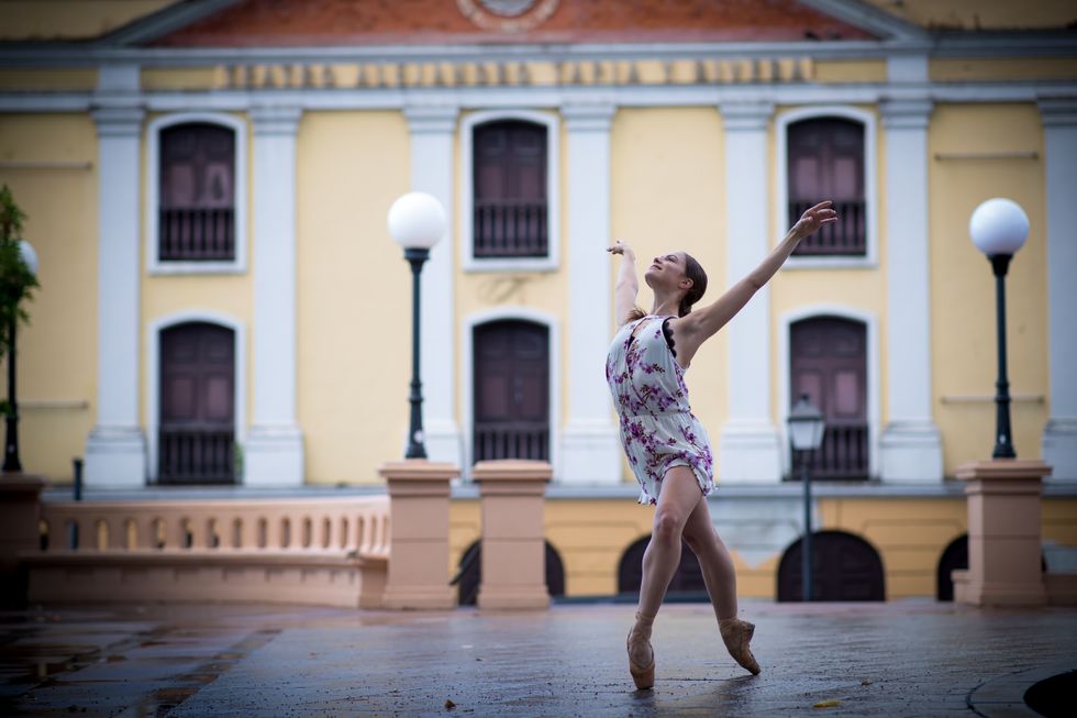 Wearing a short, flowered dress, Sabrina Landa poses in fourth position on pointe, bending her right knee. She stands on a plaza in front of a grand yellow building.