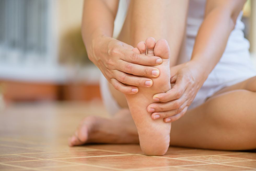 A young woman sits on the ground with her left leg tucked underneath her and grabs her right bare foot with both hands.
