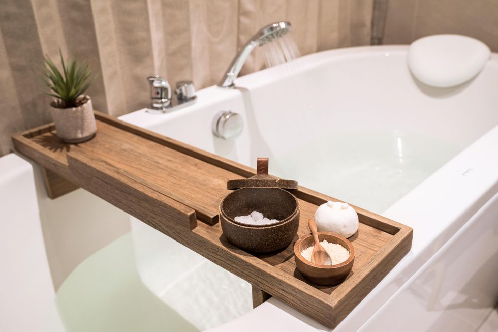A tranquil bathtub scene. A bamboo tray sits atop the tub and holds bath salts and a small, green plant.