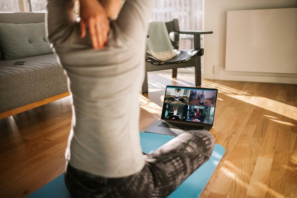 A woman in activewear, seated on a yoga mat, stretches her arm behind her head. She is seated in a living room and is doing a yoga class on her laptop with others.