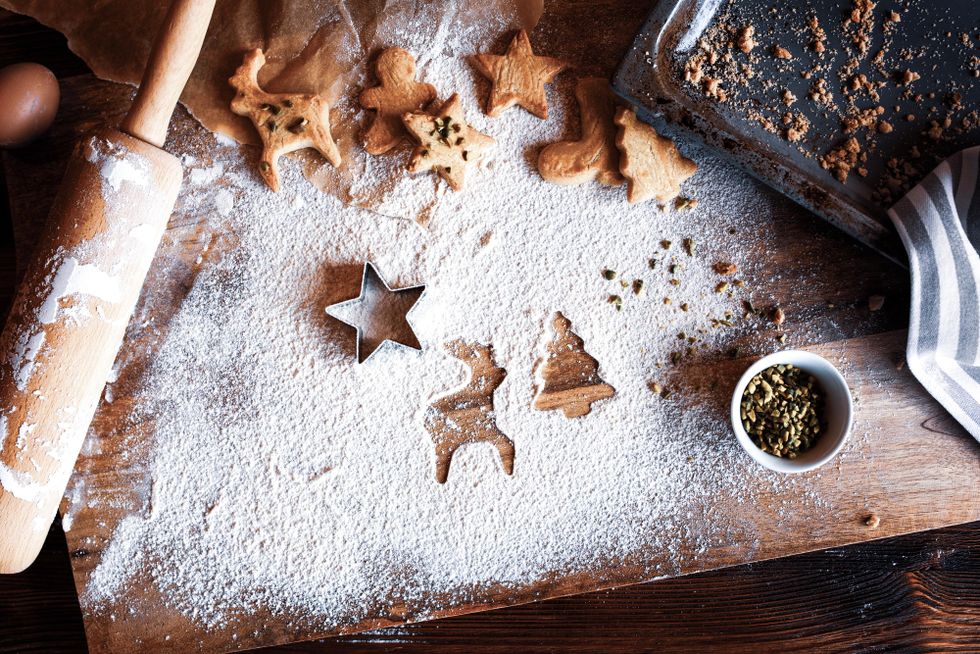 A wood table covered in flour and adorned with holiday cookie baking items, including a rolling pin, a star-shaped cookie cutter, a cookie sheet and cookies shaped like stars, reindeer and trees.