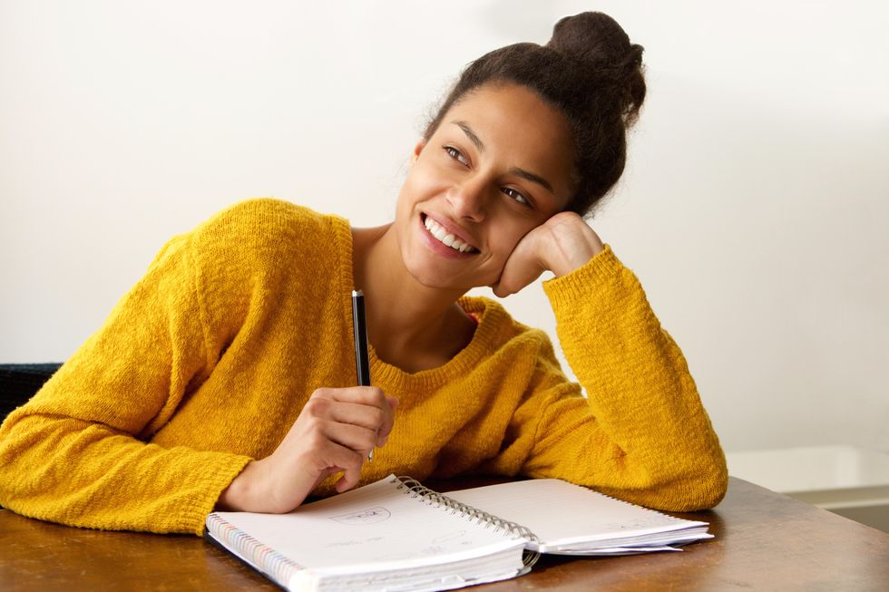 A light-skinned Black young woman, with her hair in a bun, sits thinking at a desk with a smile on her face. She is holding a pen above a notebook.